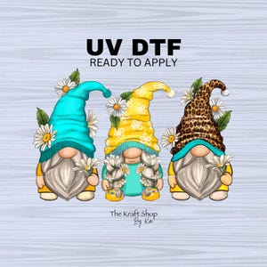 May include: Three gnomes wearing different patterned hats with flowers around their heads. The gnomes are wearing blue, yellow, and brown hats. The text "UV DTF READY TO APPLY" is at the top of the image. The text "The Kraft Shop By Kai" is at the bottom of the image.