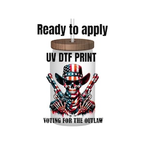 May include: A white tumbler with a wood lid and a straw. The tumbler has a skull wearing a cowboy hat with an American flag design. The text "UV DTF PRINT" and "VOTING FOR THE OUTLAW" is printed on the tumbler.