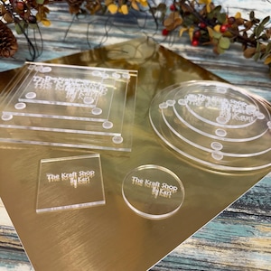 May include: A set of clear acrylic cake tiers and square bases with the text "The Kraft Shop by Kari" engraved on each piece. The pieces are arranged on a gold surface.
