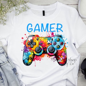 May include: White t-shirt with a colorful graphic of a video game controller and the word "GAMER" in blue text.
