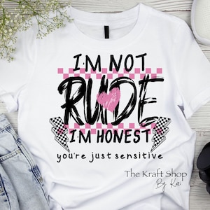 May include: White t-shirt with the text "I'M NOT RUDE I'M HONEST you're just sensitive". The word "RUDE" is in large, black, cursive letters with a pink heart in the middle. The shirt also features a pink and white checkered pattern.