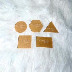 May include: A collection of five acrylic shapes: a circle, hexagon, triangle, square, and rectangle. Each shape is a light brown color and features the text "The Kraft Shop by Kari". The shapes are arranged on a white, fluffy surface.