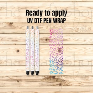 May include: Three pens featuring a gradient leopard print pattern in pink, purple, and blue, alongside a matching rectangular UV DTF pen wrap decal. The text "Ready to apply UV DTF PEN WRAP" is printed in black above the items.