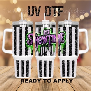 May include: Three white tumblers with black vertical stripes and silver lids. The center tumbler features the text "It's Showtime" in purple and green with a skull graphic. The words "UV DTF" and "READY TO APPLY" are also visible.