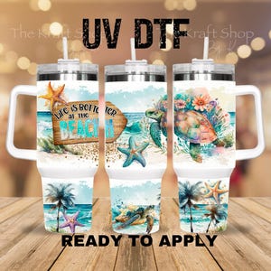 May include: Three white insulated tumblers with handles, decorated with beach-themed designs. Each features a different design: a wooden sign with text, a sea turtle, and a turtle with flowers. The text "Life is better at the beach" is visible.