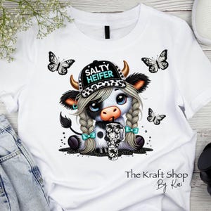 May include: White t-shirt with a cartoon cow wearing a hat that says "Salty Heifer". The cow is holding a black and white tumbler. There are butterflies on the shirt.