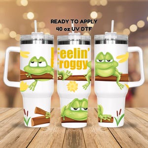 May include: Three 40 ounce stainless steel tumblers with lids and handles. The tumblers are white with a green frog graphic and the text "Feelin' Froggy".
