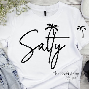 May include: White t-shirt with black text that says "Saty" in a cursive font. A palm tree graphic is above the text. The text "The Kraft Shop By Kari" is at the bottom of the shirt.