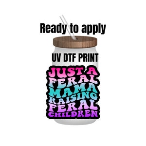 May include: A white tumbler with a wooden lid and a straw. The tumbler has a UV DTF print that says "Just a Feral Mama Raising Feral Children" in a rainbow gradient.
