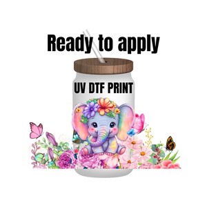 May include: A white tumbler with a wooden lid and a straw. The tumbler has a colorful illustration of a cartoon elephant wearing a flower crown and surrounded by flowers. The text "UV DTF PRINT" is printed on the tumbler.