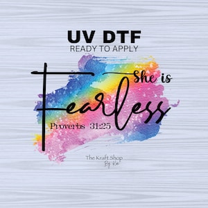 May include: A UV DTF ready to apply transfer with the words "She is fearless" in black script over a watercolor rainbow design. The text "Proverbs 31:25" is below the word "fearless". The Kraft Shop By Kai is at the bottom.