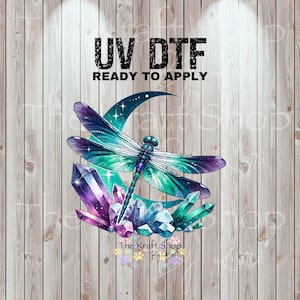 May include: A colorful UV DTF transfer design featuring a dragonfly with teal and purple wings, a crescent moon, and crystals. The text "UV DTF READY TO APPLY" is displayed at the top. The background is a wood grain pattern.