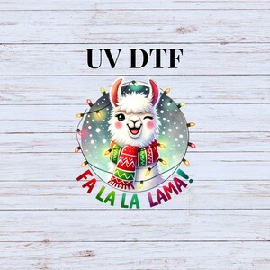 May include: A white llama with a red and green scarf is surrounded by a string of Christmas lights. The text "Fa La La Lama!" is written below the llama. The text "UV DTF" is written above the llama.