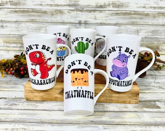 Könnte beinhalten: Satz von sechs weißen Keramikbechern mit schwarzem Text und bunten Comic-Illustrationen. Jeder Becher trägt den Text "Don't be a..." gefolgt von einem anderen humorvollen Wort. Die Wörter sind "Triceratops", "Thatwaffle", "Hippopotamus", "Cactus", "Einhorn" und "Cupcake".