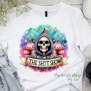 May include: White t-shirt featuring a colorful graphic. The design includes a skull in a hooded robe, circus tents, and the text "WELCOME TO THE SH*T SHOW" on a banner. The design has a whimsical, yet dark theme.