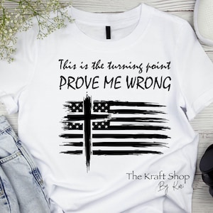 May include: White t-shirt with the text "This is the turning point PROVE ME WRONG" in black. The design includes a black cross over a stylized American flag. The Kraft Shop by Kari logo is at the bottom.