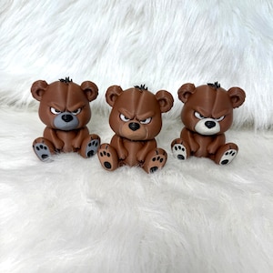 May include: Three brown bear figurines with an angry expression. Each bear has a dark brown body, black claws, and a small tuft of black hair. The bears are sitting on a white, fluffy surface.