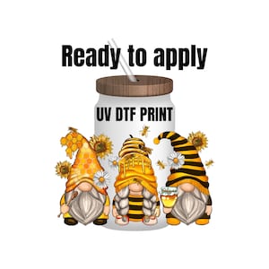 May include: Three gnomes wearing bee-themed hats and holding honey jars. The gnomes are standing in front of a white tumbler with a wooden lid and the text "UV DTF PRINT". The text "Ready to apply" is above the tumbler.