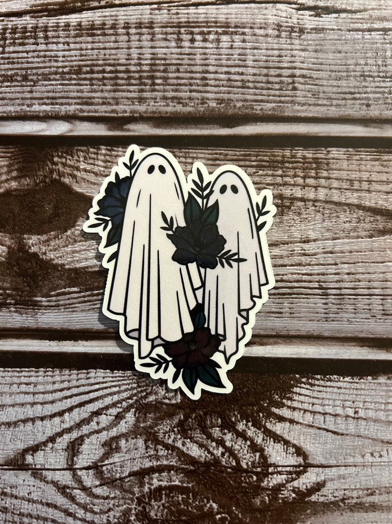 May include: Two white ghosts with black eyes and mouths, standing side by side, surrounded by dark green leaves and black flowers.