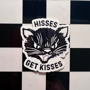 May include: Black and white sticker of a cat's face with the text "HISSES GET KISSES" on a black and white checkered background.