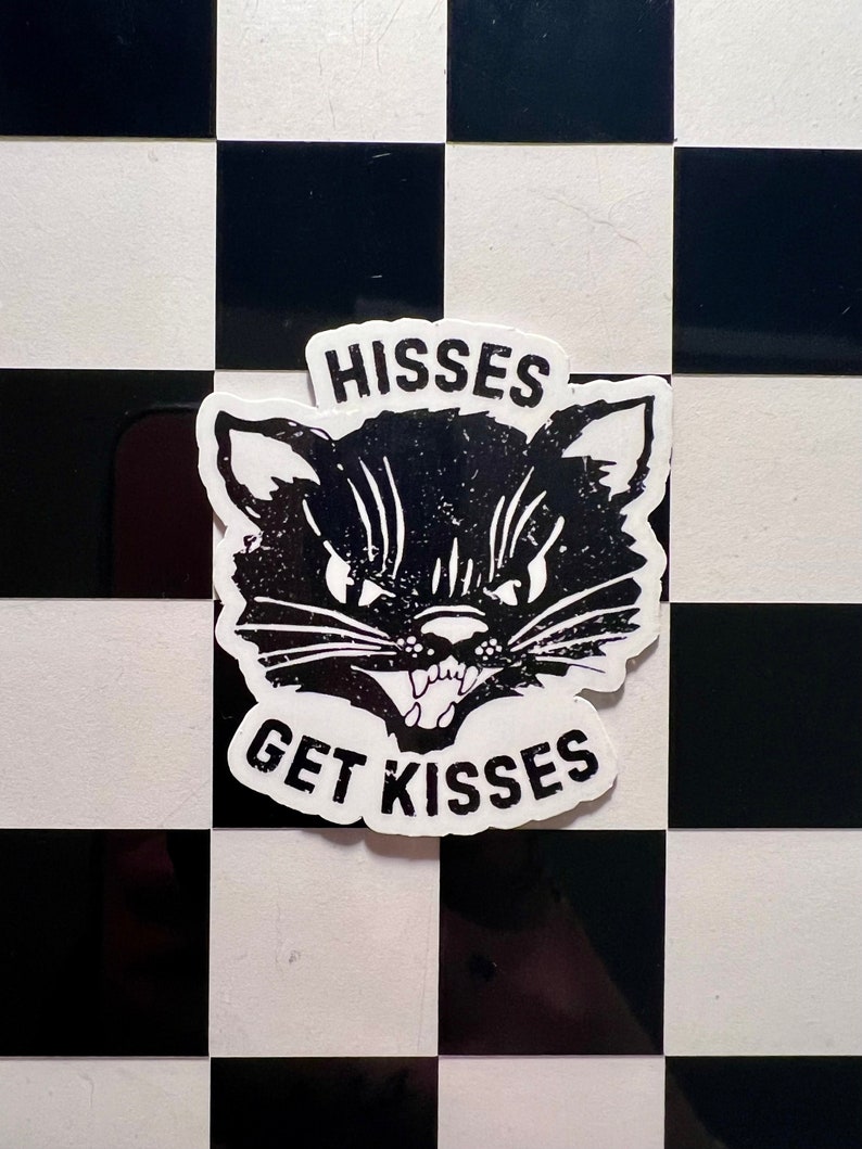 May include: Black and white sticker of a cat's face with the text "HISSES GET KISSES" on a black and white checkered background.