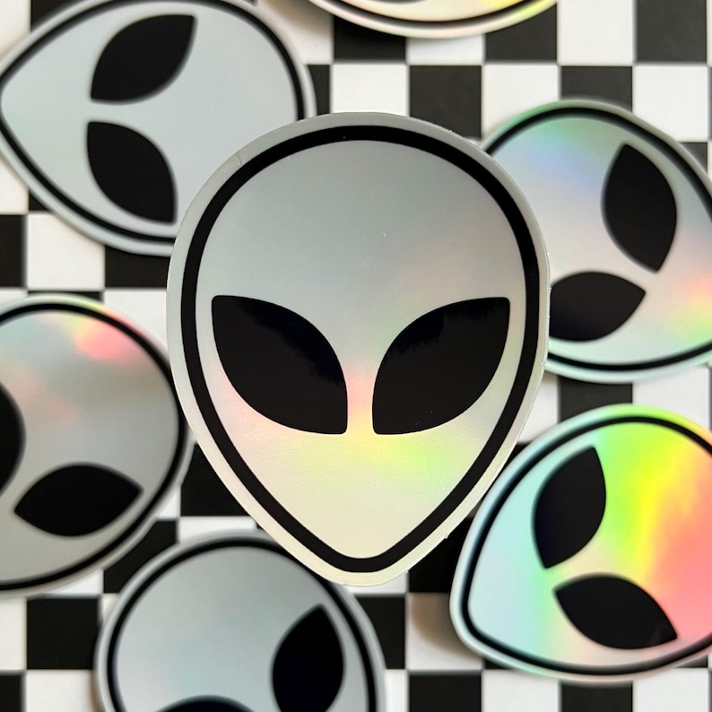 May include: A set of holographic alien head stickers with black eyes and a silver background. The stickers are arranged on a black and white checkered surface.
