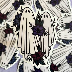 May include: Two white ghost figures with black outlines, holding hands and surrounded by purple and blue flowers. The ghosts are wearing white gowns with ruffled edges. The flowers have dark green stems and leaves.