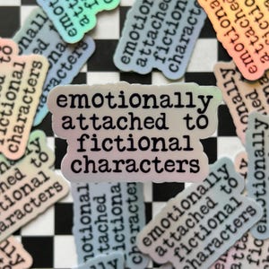May include: A holographic sticker with the text "emotionally attached to fictional characters" in black lettering. The sticker has a rainbow iridescent background.