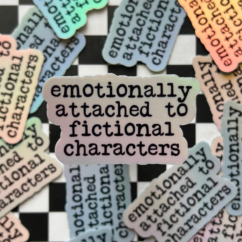 May include: A holographic sticker with the text "emotionally attached to fictional characters" in black lettering. The sticker has a rainbow iridescent background.