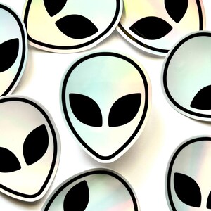 May include: A set of eight iridescent holographic stickers featuring a black alien head silhouette with two large eyes. The stickers are circular and have a black outline.