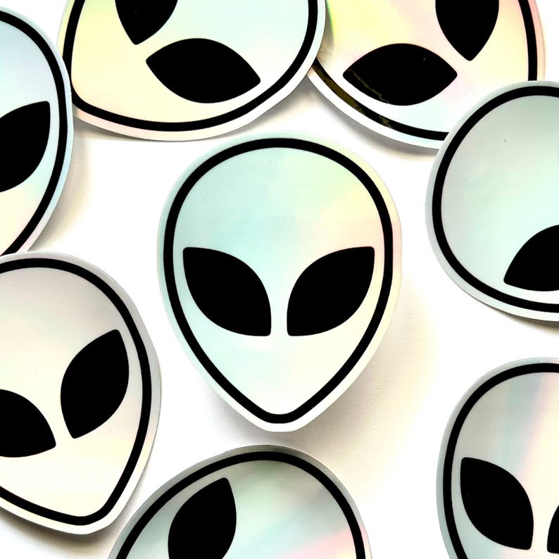 May include: A set of eight iridescent holographic stickers featuring a black alien head silhouette with two large eyes. The stickers are circular and have a black outline.