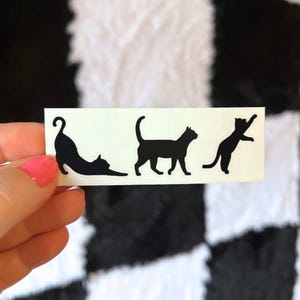 May include: A white rectangular sticker featuring three black cat silhouettes in different poses: stretching, walking, and reaching up. The sticker is held against a black and white checkered background.