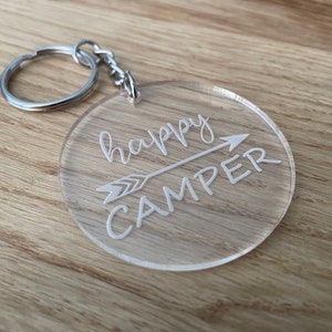 Könnte beinhalten: Ein transparenter Acryl-Schlüsselanhänger mit einem weißen Pfeil und dem Text "happy camper", der darauf graviert ist. Der Schlüsselanhänger hat einen silbernen Metallring.