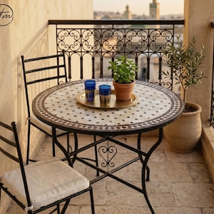 Può includere: Un tavolo rotondo in mosaico con un motivo geometrico bianco e marrone, sostenuto da una struttura in metallo nero. Due sedie in metallo nero con cuscini bianchi accompagnano il tavolo. Un vassoio dorato contiene due bicchieri blu e una pianta.