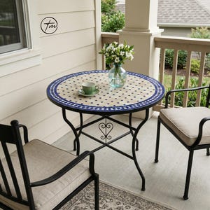 May include: A round outdoor bistro table with a mosaic tile top in beige and blue. The table has a black metal frame and is surrounded by two black metal chairs with beige cushions. A green teacup and saucer sit on the table, next to a vase of white flowers.