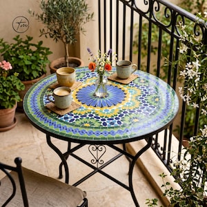 Puede incluir: Una mesa redonda de mosaico con base de metal negro. La mesa presenta un colorido patrón geométrico en tonos azules, verdes y amarillos. Dos tazas de café están sobre la mesa, junto con un pequeño jarrón con flores. La mesa tiene unos 61 cm de diámetro.