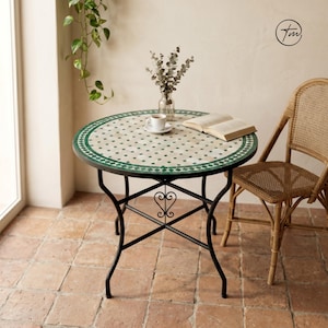 May include: A round mosaic tile table with a black metal frame. The tabletop features a white and green tile pattern. A cup of coffee and an open book rest on the table. The table is approximately 28 inches in diameter.