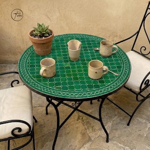 May include: A round outdoor table with a green mosaic top, surrounded by black metal chairs. The table holds four ceramic mugs, a small plant in a terracotta pot, and a paper coffee filter. The table is set on a stone patio.