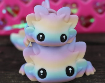 May include: A stack of two pastel-colored fantasy creatures with large black eyes. The creatures are in shades of yellow, blue, and purple. The background is a pink heart-shaped design.