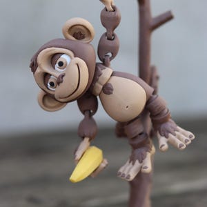 May include: A brown monkey figurine holding a yellow banana, hanging from a brown tree branch. The monkey has a playful expression and is made of a textured material.