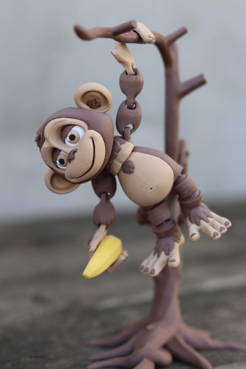 May include: A brown monkey figurine holding a yellow banana, hanging from a brown tree branch. The monkey has a playful expression and is made of a textured material.