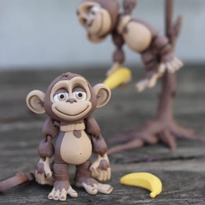 May include: Two brown monkey figurines, one hanging from a tree branch and the other standing on a wooden surface. Both monkeys have a friendly expression and are holding yellow bananas.