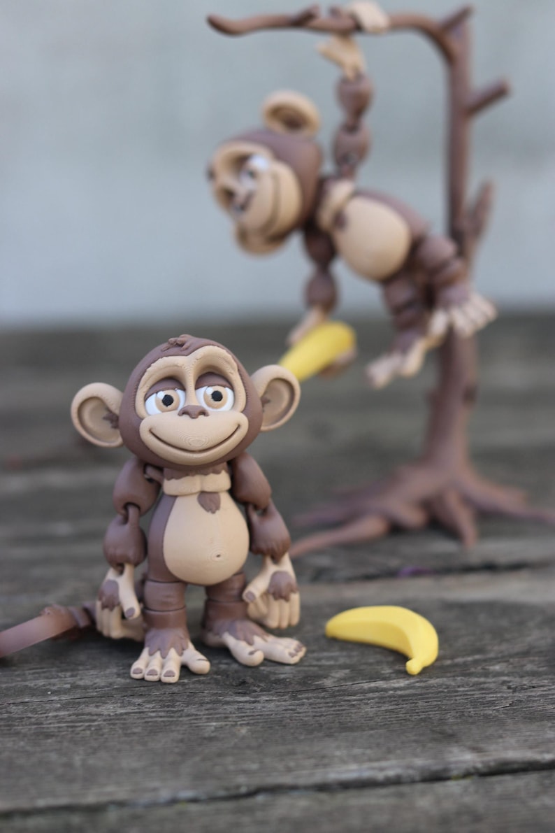 May include: Two brown monkey figurines, one hanging from a tree branch and the other standing on a wooden surface. Both monkeys have a friendly expression and are holding yellow bananas.