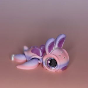 May include: A whimsical, gradient-colored bunny figurine in shades of pink, purple, and white. The bunny is lying down, with its large eye and ears visible. The figurine has a heart-shaped pupil and small heart details.