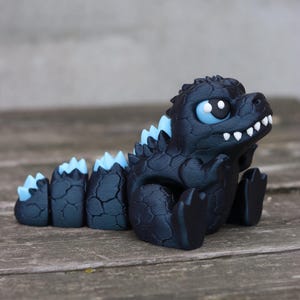 May include: A small, articulated figurine of a dark blue, reptilian creature with a textured, cracked skin pattern. It features light blue dorsal fins along its back and tail, large light blue eyes with black pupils, and small white teeth.