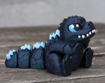 May include: A handcrafted, dark blue figurine of a cartoon-style dinosaur. The dinosaur has light blue spines, large blue eyes, and white teeth. The figurine is sitting on a wooden surface.