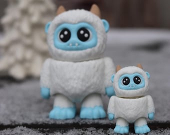 May include: Two white fuzzy Yeti figurines with blue faces and feet. The larger Yeti is standing behind the smaller Yeti.