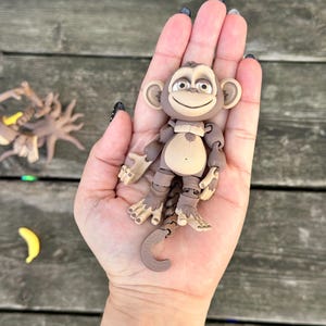 May include: A brown articulated monkey toy with a smiling face and a long tail. The monkey is posed with its arms outstretched and its legs bent. The toy is made of plastic and has a realistic appearance.