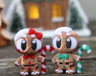 May include: Two gingerbread-style figurines with white icing hair, large eyes, and candy cane accessories. One has a red bow, the other a green bow tie. A miniature gingerbread house is in the background.