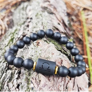 May include: A stretch bracelet featuring matte black spherical beads and a central cylindrical black bead engraved with the Libra zodiac symbol. The central bead is flanked by two small, polished gold-tone metal spacer rings.